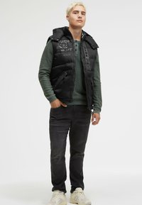 Schwarze gepolsterte Weste mit hohem Kragen, verstärkter Vorderseite und Logo-Details, getragen über einem strukturierten grünen Langarmoberteil und dunklen Jeans.