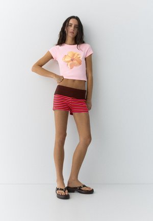 Femme debout contre un mur blanc, portant un crop top rose avec une fleur jaune, un short rayé rouge et rose, et des sandales à plateforme noires.