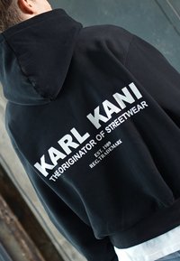 Black hoodie made of cotton. Features large white text on the back: "KARL KANI THE ORIGINATOR OF STREETWEAR EST. 1989." Simple design.