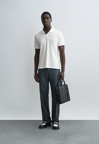 Homme portant un polo blanc à manches courtes, un pantalon gris, des mocassins noirs et blancs, tenant une mallette en cuir noir devant un fond uni.