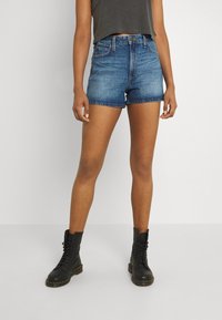 Persona que lleva puestos unos pantalones cortos de mezclilla azul de cintura alta, botas negras con cordones y una camiseta sin mangas oscura de corte cropped, de pie contra un fondo liso.