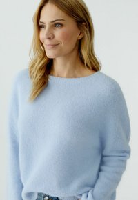 Frau mit blondem Haar, die einen hellblauen, weich gestrickten Pullover trägt und mit einem leichten Lächeln zur Seite schaut vor einem einfarbigen Hintergrund.