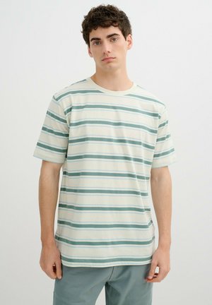 Joven con camiseta blanca de manga corta con rayas horizontales verdes y pantalones azul claro, de pie frente a un fondo liso.