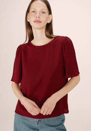 Blouse bordeaux à manches courtes bouffantes, col rond et tissu texturé, associée à un jean en denim clair. Design minimaliste, finition lisse.