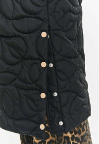 Čierna prešívaná látka s veľkými vzormi listov, upevnená béžovými a striebornými cvočkami, v páre s odevom s leopardím vzorom pod ňou.