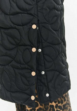 Černá prošívaná látka s velkými vzory listů, zajištěná béžovými a stříbrnými cvočky, doplněná o oděv s leopardím potiskem pod ní.