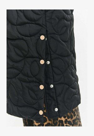 Czarna pikowana tkanina z dużymi wzorami liści, zapinana na beżowe i srebrne zatrzaski, zestawiona z ubraniem w panterkę pod spodem.