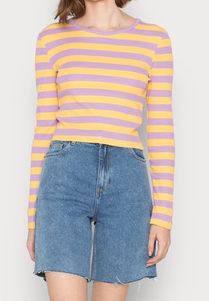 Kvinde iført en langærmet gul og lavendelstribet top og højtaljede blå denimshorts med rå kanter.