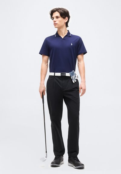 Νεαρός άνδρας με μπλε πόλο και μαύρο παντελόνι κρατώντας ένα γκολφ κλαμπ, με γάντι γκολφ κρυμμένο στη λευκή ζώνη, πάνω σε απλό φόντο.