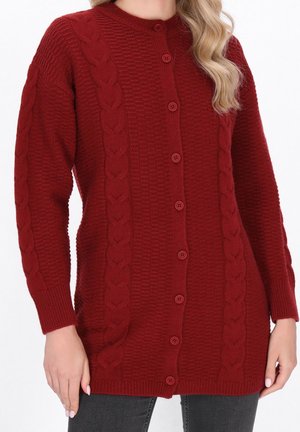 Kobieta w czerwonym, długim, zapinanym na guziki swetrze na drutach z warkoczowym wzorem, z prążkowanymi ściągaczami na rękawach i dole, nosząca ciemne jeansy.