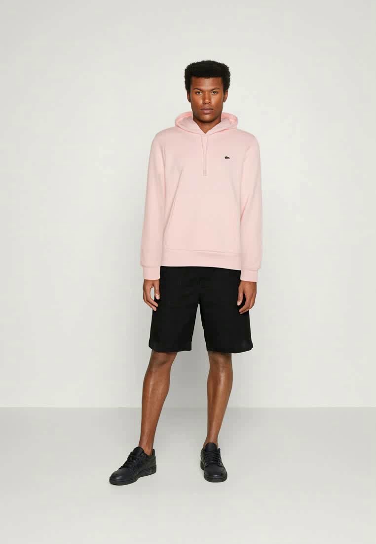 Haut à capuche rose clair avec une fermeture éclair à l'avant et un ourlet côtelé, associé à un short noir et des baskets noires ; design minimaliste avec un accent logo.