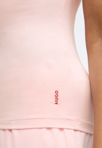 Lichtroze tanktop van gladde stof, met een rood "HUGO" logo vlakbij de zoom. Ontworpen met een aangesloten silhouet en een ronde halslijn.