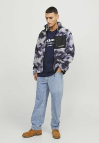 Chaqueta de forro polar en un patrón de teñido anudado en púrpura y blanco, con cierre de cremallera y un bolsillo en el pecho negro. Lleva puesta una camisa azul marino y jeans claros.