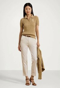 Γυναίκα που φοράει καφέ πλεκτό πόλο, εκρού παντελόνια, κρατάει το ίδιο πουλόβερ και καφέ τακούνια, στηρίζεται σε απλό φόντο.