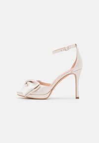 White satin high-heeled sandal with a front knot detail, open toe, ankle strap, and a slim stiletto heel. Smooth texture with a minimalist design.