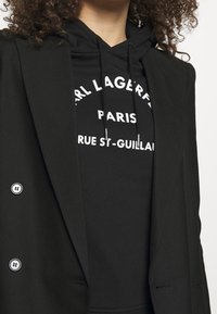 Mujer vestida con un blazer negro sobre una sudadera negra con texto blanco que dice "Karl Lagerfeld Paris Rue St-Guillaume."