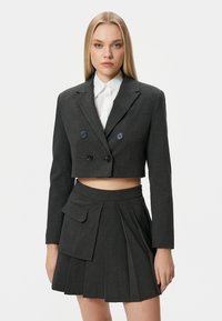 Gekorte donkergrijze blazer met dubbele knopen, gecombineerd met een geplooide rok met een opgestikte zak, gedragen over een witte blouse met knopen.