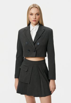 Gekorte donkergrijze blazer met dubbele knopen, gecombineerd met een geplooide rok met een opgestikte zak, gedragen over een witte blouse met knopen.