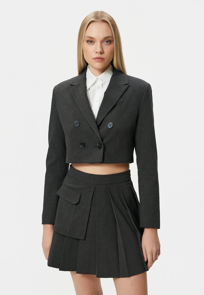 Gekorte donkergrijze blazer met dubbele knopen, gecombineerd met een geplooide rok met een opgestikte zak, gedragen over een witte blouse met knopen.