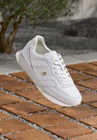 Hvit lær sneakers med snøring og gulllogo detalj, svevende over tre firkantfliser med små grå steiner i en utendørs setting.
