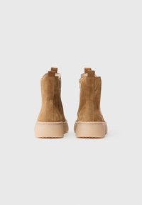 Une paire de bottines en suède beige avec fermeture éclair latérale et semelles en caoutchouc beige, vues de l'arrière sur un fond blanc.