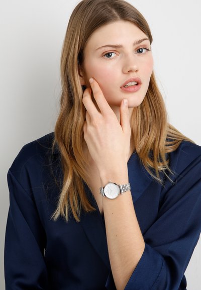 Silberne Armbanduhr mit rundem Ziffernblatt, metallischem Mesh-Armband, strukturiertem Zifferblattdesign, getragen an einer Hand vor einem neutralen Hintergrund.