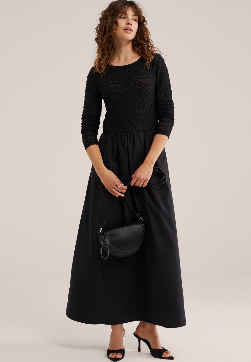 Femme aux cheveux ondulés portant une longue robe noire à manches texturées, tenant un petit sac à main noir et portant des talons ouverts noirs.
