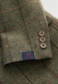 Ullherringbone-blazer med ett grönt och brunt mönster, tre bruna knappar och en broderad marinblå etikett med röd text.