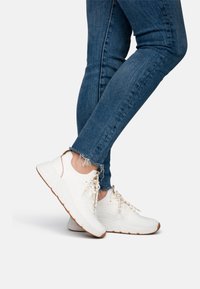 Zapatillas de punto blancas con patrones texturizados, cordones beige y suela de goma marrón claro; combinadas con unos jeans de mezclilla azules deshilachados.