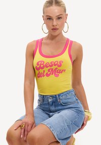 Femme aux cheveux blonds portant un débardeur jaune avec une bordure rose et le texte "Besos del Mar,", portant un short en jean, des boucles d'oreilles créoles en or et des accessoires dorés.