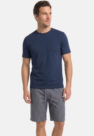 Mężczyzna ubrany w granatowy t-shirt z krótkim rękawem i szare, kraciaste szorty do kolan, stoi na jednolitym białym tle.
