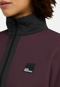 Fleecejack in donker bordeaux met een hoge hals. Voorzien van een zwarte rits en een zwart merkembleem op de borst. Glad, textuurstof.