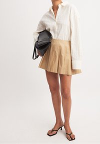 Chemise blanche à manches longues et à boutons, avec de subtils rayures verticales, associée à une jupe plissée beige et des sandales à talons noires ; tenant un sac noir.