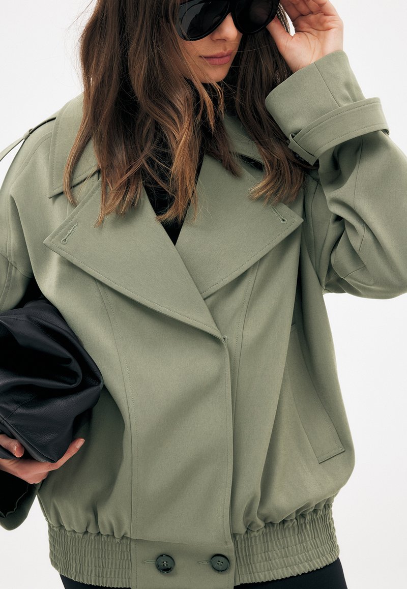 Kvinde iført en oversized lysegrøn jakke og store sorte solbriller, der holder en sort læder clutch taske med løst bølget hår.