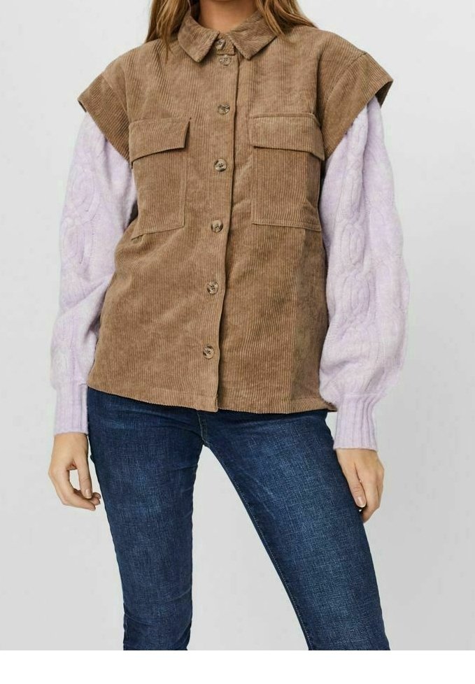 Femme portant un gilet sans manches en velours côtelé marron boutonné sur un pull en tricot torsadé lavande et un jean bleu foncé.