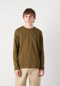 Langärmliges, olivgrünes T-Shirt aus weicher Baumwolle mit rundem Ausschnitt und einem kleinen quadratischen Logopatch auf der Brust. Glatte Textur, entspannter Schnitt.