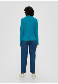 Turkusowa marynarka z kołnierzykiem, długimi rękawami i luźnym krojem, zestawiona z luźnymi niebieskimi dżinsami i białymi trampkami.