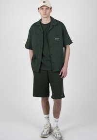 Jeune homme portant une chemise vert foncé à manches courtes boutonnée, un short assorti, des chaussettes blanches avec logos, des baskets grisées et une casquette de baseball beige.