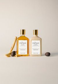 Deux flacons à bouchons dorés de shampooing et après-shampooing Rahua avec des bâtonnets en bois naturel et des graines foncées sur un fond clair.