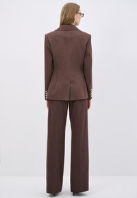 Completo marrone su misura caratterizzato da un blazer aderente con uno spacco sul retro e pantaloni a gamba ampia; progettato con bottoni dorati come dettaglio.