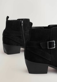 Botines de ante negro con un tacón bajo tipo bloque, que cuentan con una cremallera lateral, costuras decorativas y una correa con un detalle de hebilla plateada.