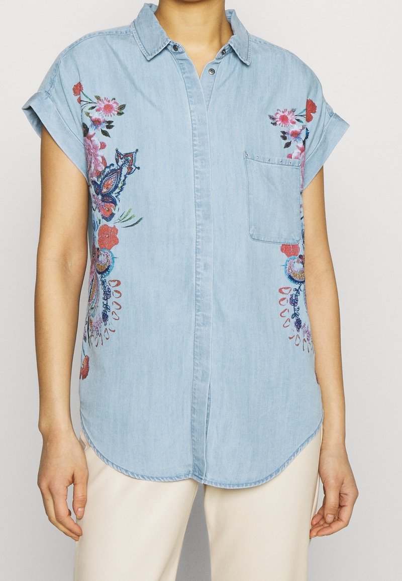 Camicia di jeans blu chiaro con maniche corte arrotolate, caratterizzata da un ricamo floreale colorato e una tasca a toppa sul lato sinistro.