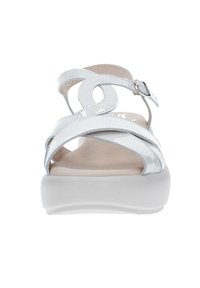 Sandalias de charol blanco con tiras entrecruzadas y un acento circular, que presentan un tacón cuña bajo y cierre de hebilla ajustable.