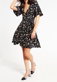 Mujer vestida con un vestido wrap negro de flores con mangas cortas y zapatos de tacón bloque con correas al tobillo, de pie frente a un fondo blanco.