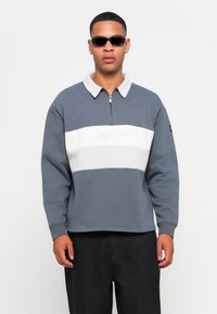 Grå sweatshirt med en hvid vandret stribe, lynlås krave og broderet tekst. Båret med sorte bukser og ovale solbriller.