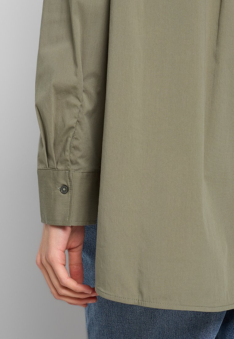 Langärmliges, olivgrünes Hemd aus glattem, weichem Stoff. Die Ärmelbündchen verfügen über eine Knopfdetail. Kombiniert mit blauer Jeans an der Taille.