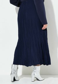 Gonna maxi plissée blu navy in tessuto testurizzato, abbinata a stivali alla caviglia argentati lucidi con tacco a blocco e zip laterale.