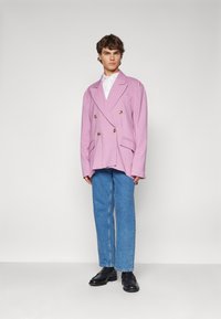 Dobbeltspent rosa blazer med gullknapper, kombinert med lyseblå rette jeans og sorte sko. Bakgrunnen er enkel og lys.