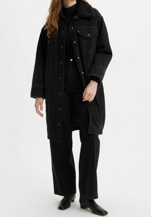 Femme portant un long manteau en denim noir avec un col en mouton retourné, pantalon noir, haut noir et bottines noires à talons, debout sur un fond blanc.