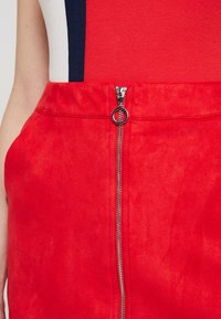 Jupe en daim rouge avec une fermeture éclair à l'avant munie d'un anneau circulaire. Le tissu présente une texture lisse et une coloration uniforme.